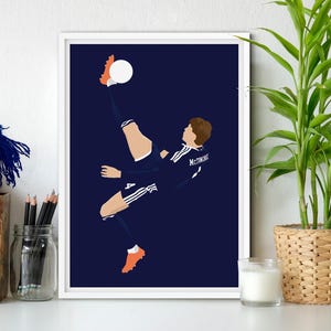 May include: A framed print showing a soccer player mid-kick, with a white ball. The player wears a navy blue kit with white stripes and orange boots, set against a dark blue backdrop. The print is displayed on a white surface, with a plant in a basket.