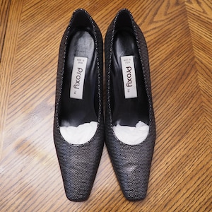 May include: A pair of black and silver pumps with a textured herringbone pattern. The interior features a white label with the text Proxy TM and Made in Spain. The shoes have a pointed toe and a black lining, with white paper stuffing inside.
