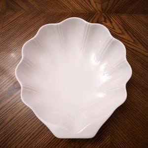 May include: A white ceramic bowl shaped like a large seashell. The bowl features a scalloped edge and embossed vertical lines radiating from the center toward the rim, creating a textured surface that mimics the appearance of a natural shell.