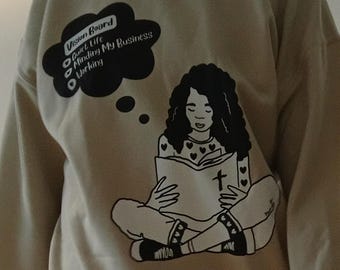 以下が含まれることがあります： ベージュの長袖スウェットシャツ。表紙に十字架が描かれた本を読む人物の黒いグラフィックがプリントされています。吹き出しには「Vision Board, Quiet Life, Minding My Business, Working」というテキストがあり、袖には「1 Thessalonians 4:11」とプリントされています。