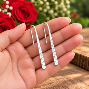 May include: A pair of silver-toned earrings featuring a long, slender, rectangular bar design with a hammered, textured finish. The earrings have a curved hook top that extends into a straight, vertical drop, creating a clean, metallic look.