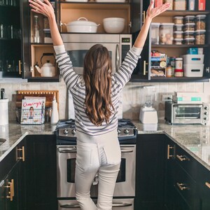 May include: A kitchen featuring dark cabinets with gold hardware, a stainless steel stove, and a microwave. Open cabinets reveal white ceramic cookware and various clear food storage containers. A cookbook titled Cravings is visible.