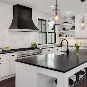 May include: A kitchen featuring white cabinetry, a large central island with a black countertop, and black bar stools. A black range hood sits above a stainless steel stove, complemented by black window frames and two glass pendant lights.