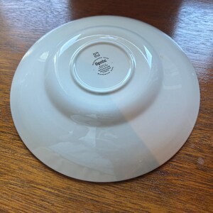 Puede incluir: Un plato de cerámica blanca visto desde la parte inferior, con un sello central que dice Spode, Made in England, Geranium, Spode Design y Microwave Safe. El plato tiene una base circular lisa y un borde ancho ligeramente curvado.