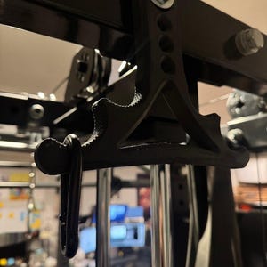 Puede incluir: Un mango de sujeción para gimnasio en forma de T, impreso en 3D en color negro con superficie texturizada, que presenta tres orificios circulares a lo largo del vástago vertical y un mosquetón de metal sujeto a un lado, montado en una máquina.