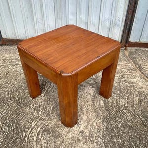 Puede incluir: Una mesa auxiliar cuadrada de madera con un acabado liso en color marrón medio. La mesa presenta una superficie plana con esquinas ligeramente redondeadas y cuatro patas rectas y robustas que ofrecen una base estable.