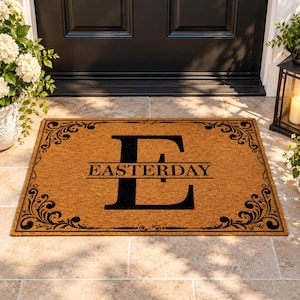 May include: A rectangular tan coir doormat featuring a large black letter E in the center with the name EASTERDAY printed horizontally across it. The mat is decorated with a black ornate scrollwork border along all four edges.