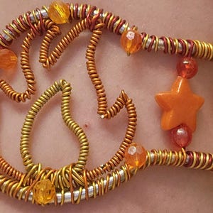 May include: A wire-wrapped cuff bracelet featuring a central flame-like design made of coiled copper-colored wire. The piece is accented with orange and yellow beads, including round spheres and star-shaped charms, integrated into the metal structure.