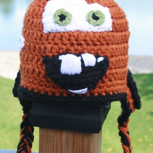 Może przedstawiać: Czapka na drutach w kształcie kreskówkowej postaci samochodu, wykonana z pomarańczowej, czarnej i białej włóczki. Czapka ma czarny brzeg i długi, warkoczowy ogon.