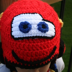 May include: Red crocheted hat with black and white details, resembling a cartoon car with large eyes. The hat has yellow accents on the sides.