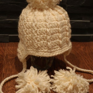 May include: Cream-colored, crocheted winter hat with ear flaps and two dangling pom-poms. The hat features a large pom-pom on top. The hat is made of a soft, textured yarn, perfect for cold weather. The hat is sitting on a wooden surface.