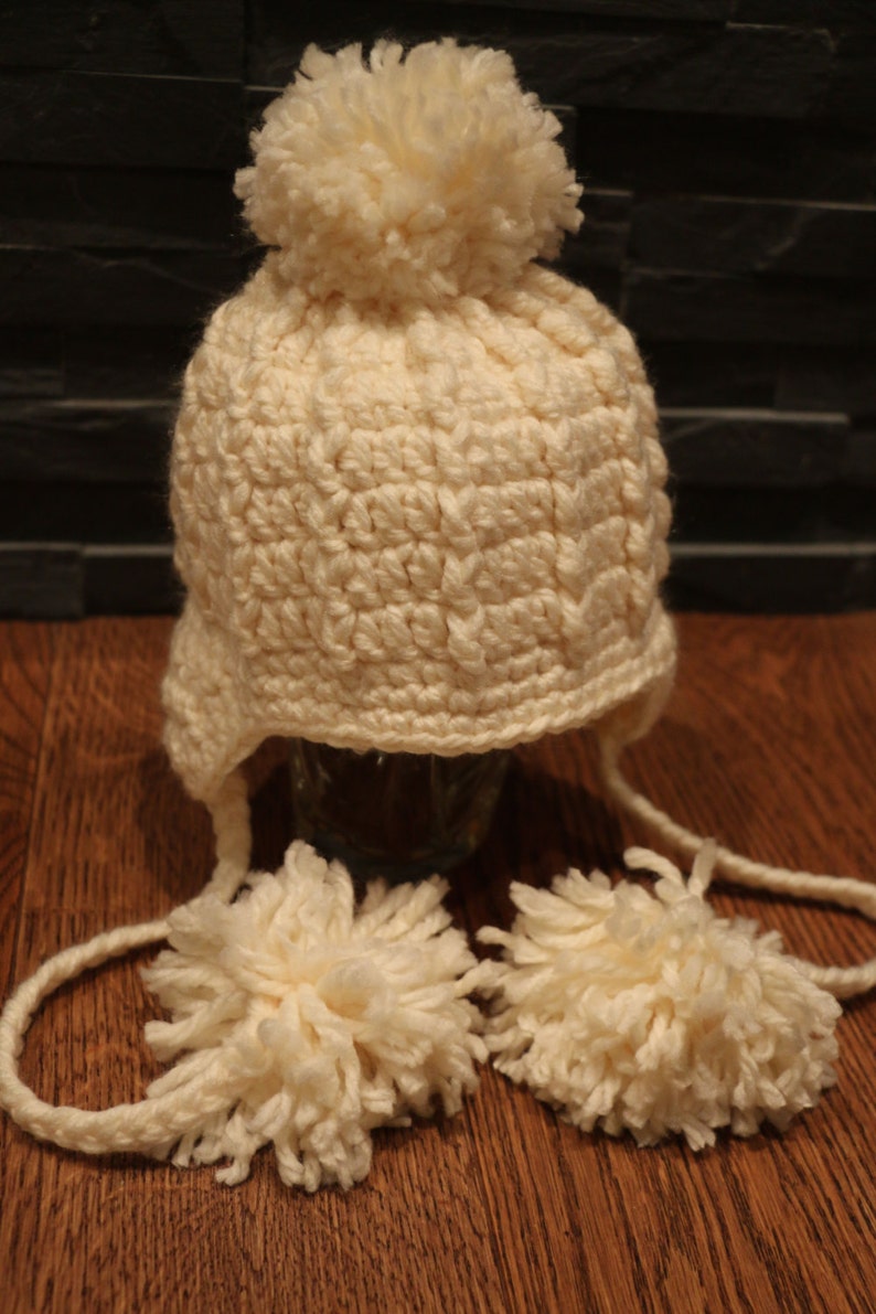 May include: Cream-colored, crocheted winter hat with ear flaps and two dangling pom-poms. The hat features a large pom-pom on top. The hat is made of a soft, textured yarn, perfect for cold weather. The hat is sitting on a wooden surface.