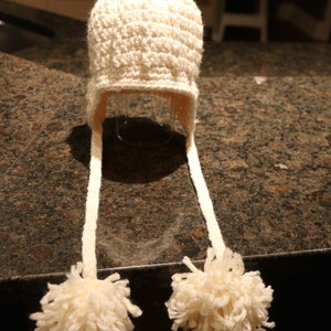 May include: Cream-colored, hand-crocheted hat with ear flaps and a large pom-pom on top. The hat has long, braided ties ending in fluffy pom-poms. The hat is made of a soft, textured yarn, perfect for keeping warm in cold weather.