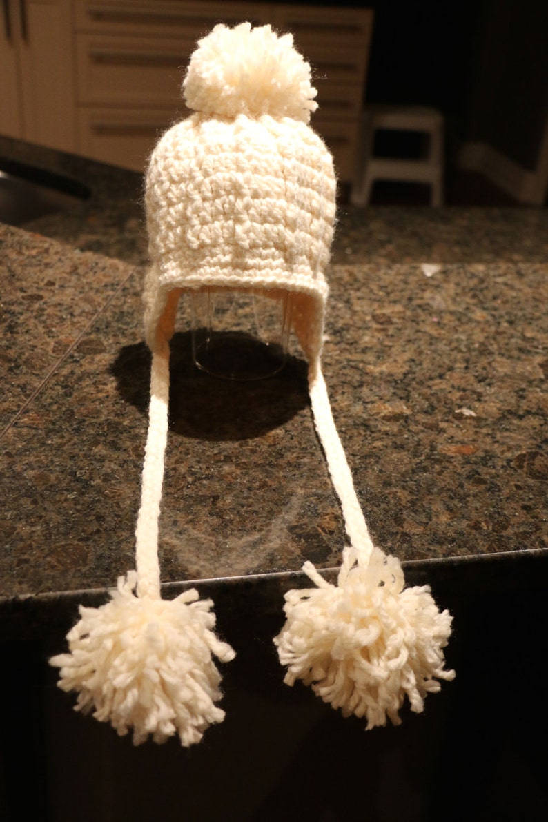 May include: Cream-colored, hand-crocheted hat with ear flaps and a large pom-pom on top. The hat has long, braided ties ending in fluffy pom-poms. The hat is made of a soft, textured yarn, perfect for keeping warm in cold weather.