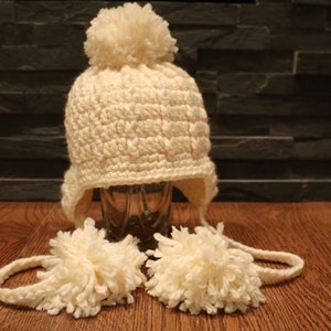May include: Cream-colored, crocheted winter hat with ear flaps and a large pom-pom on top. Two matching pom-poms are attached to the hat with a crocheted cord. The hat is displayed on a wooden surface.