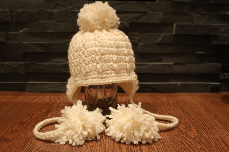 May include: Cream-colored, crocheted winter hat with ear flaps and a large pom-pom on top. Two matching pom-poms are attached to the hat with a crocheted cord. The hat is displayed on a wooden surface.