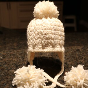May include: Cream-colored, crocheted winter hat with ear flaps and two fluffy pom-poms. The hat has a large pom-pom on top and ties under the chin. A cozy, handmade accessory for cold weather.