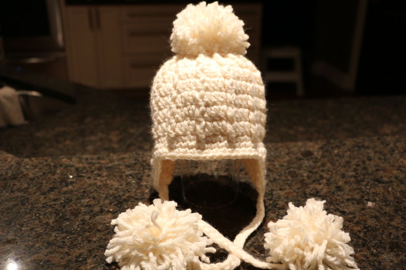 May include: Cream-colored, crocheted winter hat with ear flaps and two fluffy pom-poms. The hat has a large pom-pom on top and ties under the chin. A cozy, handmade accessory for cold weather.