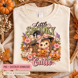 May include: A cream-colored t-shirt featuring a graphic of a hedgehog wearing a witch hat, surrounded by pumpkins, flowers, and autumn leaves. The text reads Little Spooky Cutie. The design includes pink bows, a lantern, and small mushrooms.