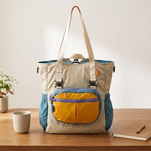 May include: A beige tote bag featuring a large mustard yellow front pocket with a purple zipper, teal side panels, and long beige shoulder straps. The bag includes black hardware, a small orange brand tag, and a top opening with a zipper closure.