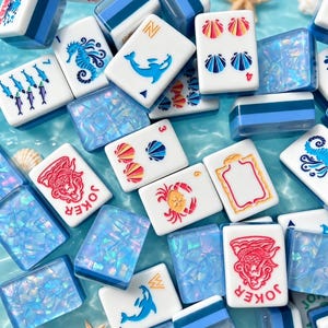 May include: A collection of rectangular mahjong tiles featuring white faces with blue, red, and orange illustrations of sea creatures, shells, and joker text, interspersed with translucent blue iridescent tiles on a water-like surface.