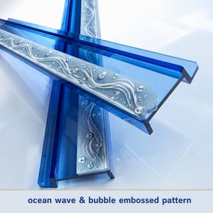 Puede incluir: Dos estantes de vidrio azul en forma de L con un patrón blanco esmerilado en relieve de olas oceánicas y burbujas. El texto ocean wave & bubble embossed pattern está impreso en la parte inferior, describiendo el diseño.