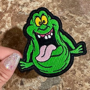 May include: Green and black embroidered patch of a cartoon ghost with a large mouth, sticking out its tongue. The ghost has yellow eyes and a toothy grin.