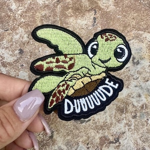 May include: An embroidered patch featuring a light green cartoon sea turtle with brown spots on its flippers and shell. The turtle has large black and white eyes and a smiling mouth. Below the turtle, the word DUUUUDE is stitched in white.