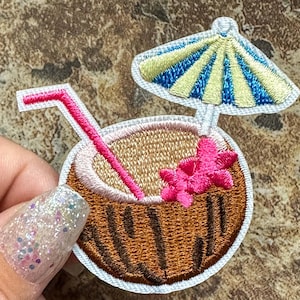 May include: Embroidered patch of a brown coconut with a pink flower, a pink straw, and a blue and yellow striped umbrella.