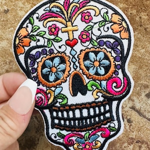 May include: A colorful embroidered sugar skull patch with floral details, a cross, and a heart. The skull is white with black eyes and teeth, and the flowers are in shades of pink, orange, yellow, green, blue, and purple.