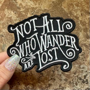 May include: Black embroidered patch with the quote "NOT ALL WHO WANDER ARE LOST" in white script. The patch has a decorative border and is likely for clothing or accessories.