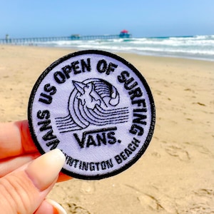 May include: A white round patch with black text and a black and white graphic of a surfer riding a wave. The text reads "Vans US Open of Surfing Huntington Beach" and "Vans".