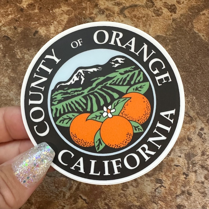 May include: A black and white sticker with the text "County of Orange California" around the edge. Inside the circle is a stylised image of a mountain range, a field of orange trees, and three oranges.