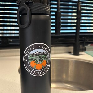 May include: Black reusable water bottle with a white logo that says "County of Orange California" and features a mountain range and three oranges.