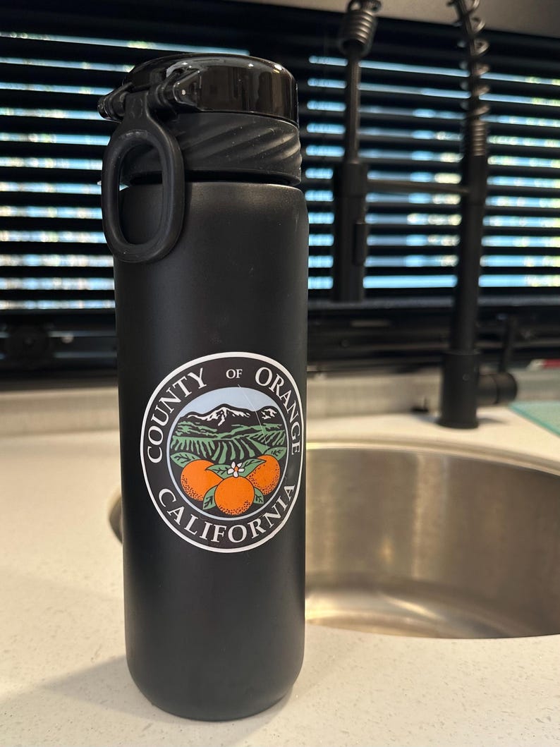 May include: Black reusable water bottle with a white logo that says "County of Orange California" and features a mountain range and three oranges.