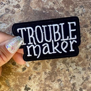 May include: A black embroidered patch with the words "TROUBLE maker" in white script. The patch has a black border and is rectangular in shape. The patch is held by a hand with glittery nail polish.