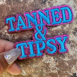 May include: A glittery pink patch with the words "TANNED & TIPSY" embroidered in bright blue. The patch has a sparkly texture and is likely designed to be sewn or ironed onto clothing or accessories.