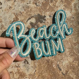 May include: A teal and white glittery patch with the words "Beach Bum" in a cursive font. The patch has a teal outline and a white interior, with a glittery texture. The patch is held by a hand.