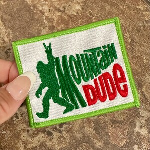 May include: Green and red embroidered patch with the text "Mountain Dude" and a green silhouette of a Bigfoot-like creature with a raised fist.