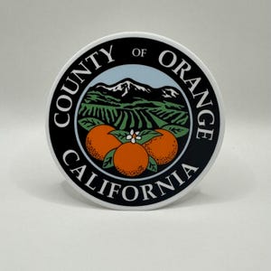 May include: Black and white sticker with the text "County of Orange California" in a circle around a stylised image of a mountain range, orange groves, and three oranges.