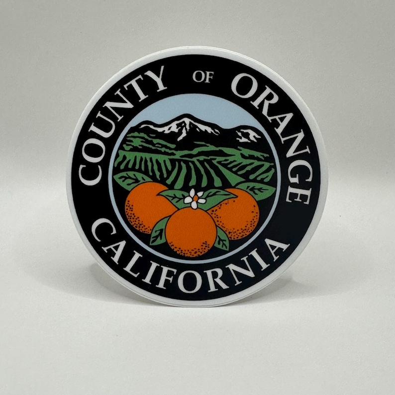 May include: Black and white sticker with the text "County of Orange California" in a circle around a stylised image of a mountain range, orange groves, and three oranges.