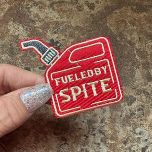 May include: A red patch in the shape of a gas can with the words "FUELED BY SPITE" embroidered in cream-colored thread. The patch has a black nozzle and a white border.