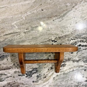 May include: A wall-mounted wooden shelf featuring a flat rectangular top surface with a grooved texture, supported by two vertical brackets connected by a horizontal rod underneath, all finished in a medium brown wood grain tone.