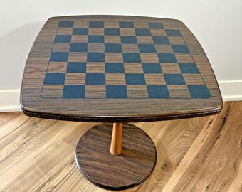 May include: A square table with a wood grain finish featuring a dark blue and brown checkered pattern on the top surface. The table is supported by a single central pedestal column that connects to a circular base with a matching wood grain.