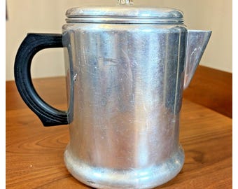 Op de afbeelding: Een zilverkleurige aluminium percolator koffiepot met een zwart hittebestendig handvat aan de zijkant, een brede cilindrische behuizing met zichtbare krassen, en een doorzichtige glazen knop op het deksel met een metalen tuit aan de rechterkant.