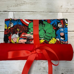 May include: A red fabric roll with a red ribbon tied around it. The fabric roll is decorated with a black and white print of various superheroes, including Captain America, Hulk, Thor, and Iron Man.