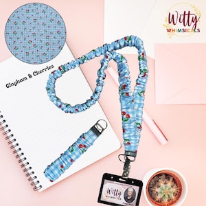 May include: A blue gingham and cherry print lanyard with a black badge holder. The lanyard is made of a soft, gathered fabric. The image also includes a notebook, pen, and a small potted cactus.