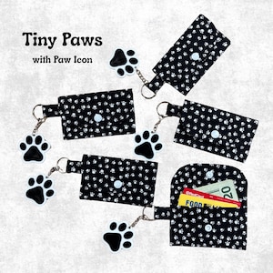 May include: Five rectangular black fabric pouches with a white paw print pattern, each featuring a snap closure and a dangling black and white paw print charm on a metal keyring. One pouch is open, revealing a twenty-dollar bill and a credit card.