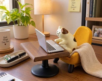 May include: A ceramic cat figurine with brown and white patches sits in a small yellow armchair, typing on a miniature silver laptop placed on a wooden desk stand with a black pedestal base. A beige knit blanket is draped over the chair.