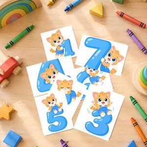 Könnte beinhalten: Ein Set aus fünf quadratischen weißen Karten mit orangefarbenen Zeichentrickkätzchen in blauen Latzhosen, die jeweils mit einer großen blauen Zahl interagieren. Dargestellt sind die Zahlen eins, drei, vier, fünf und sieben. Die Karten liegen verstreut auf einer Holzoberfläche.